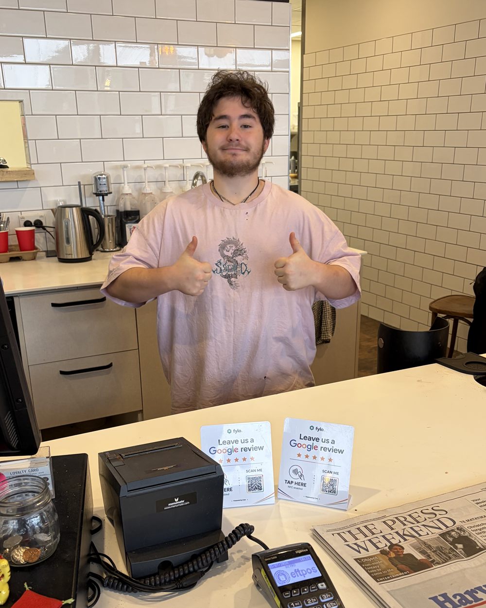 A cafe worker gives a thumbs up behind the counter, with two Fylo review stands beside the eftpos machine.