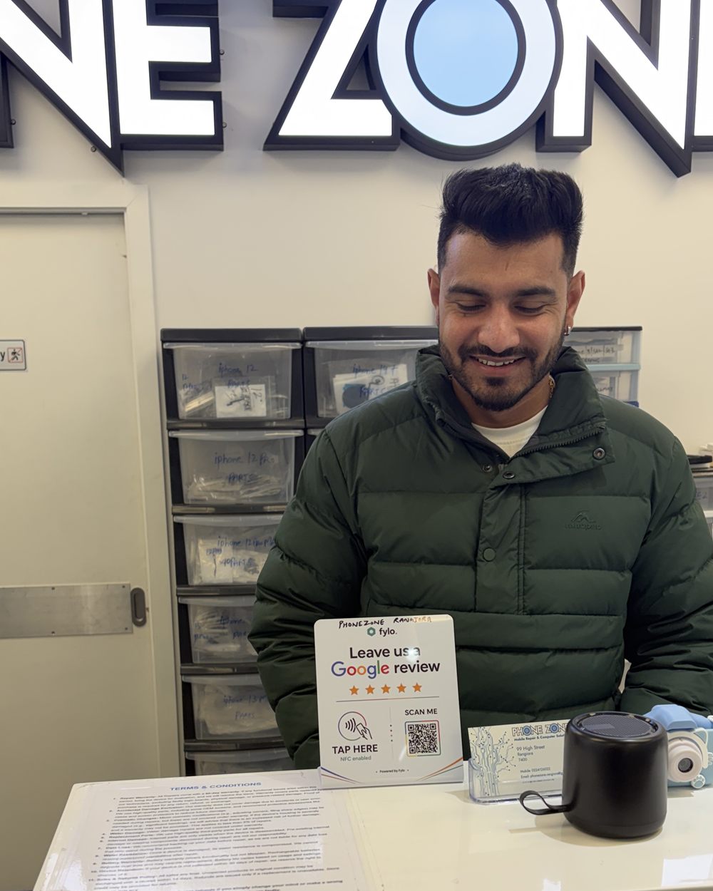 A phone repair shop owner behind the front desk with a Fylo review stand next to the till.