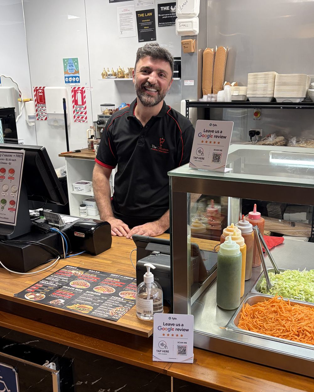 A takeaway shop owner smiles behind his counter, with Fylo review stands on the counter and the salad bar.